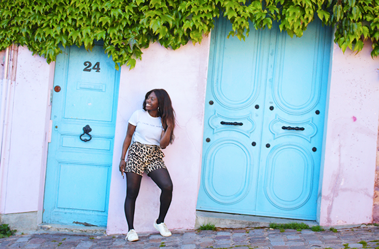 Meltingsisters-a smiling girl standing against a pink wall and two blue doors in Paris Paris is: magical Montmartre in the spring